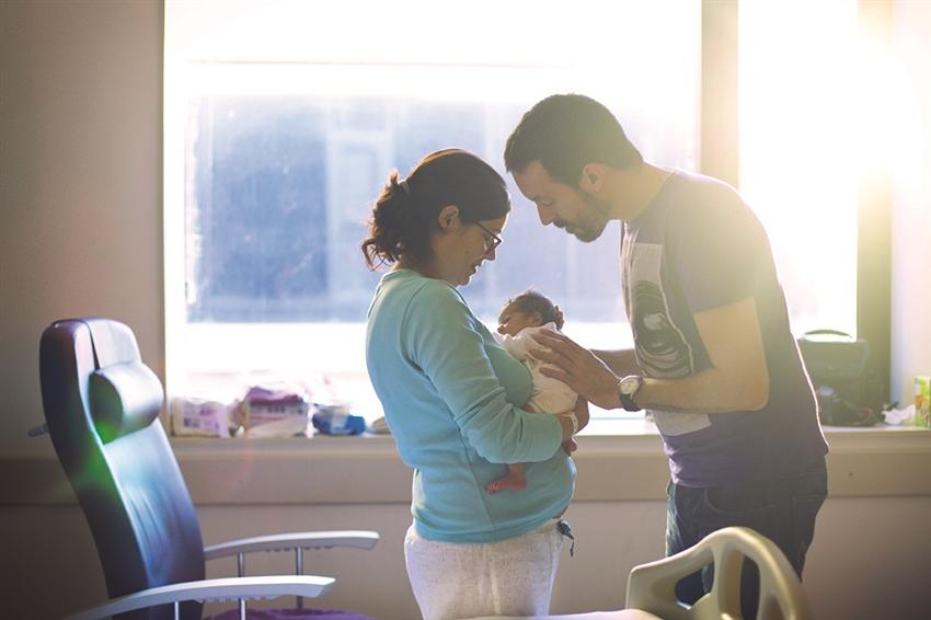 New parents standing in hospital room, mom cradling newborn with dad looking on