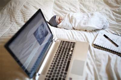 A baby laying swaddled on a bed next to a notepad with pen and a laptop