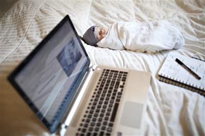 A baby laying swaddled on a bed next to a notepad with pen and a laptop A baby laying swaddled on a bed next to a notepad with pen and a laptop