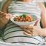 Pregnant woman reclining on a couch with a noodle bowl resting on her belly and chopsticks in her hand
