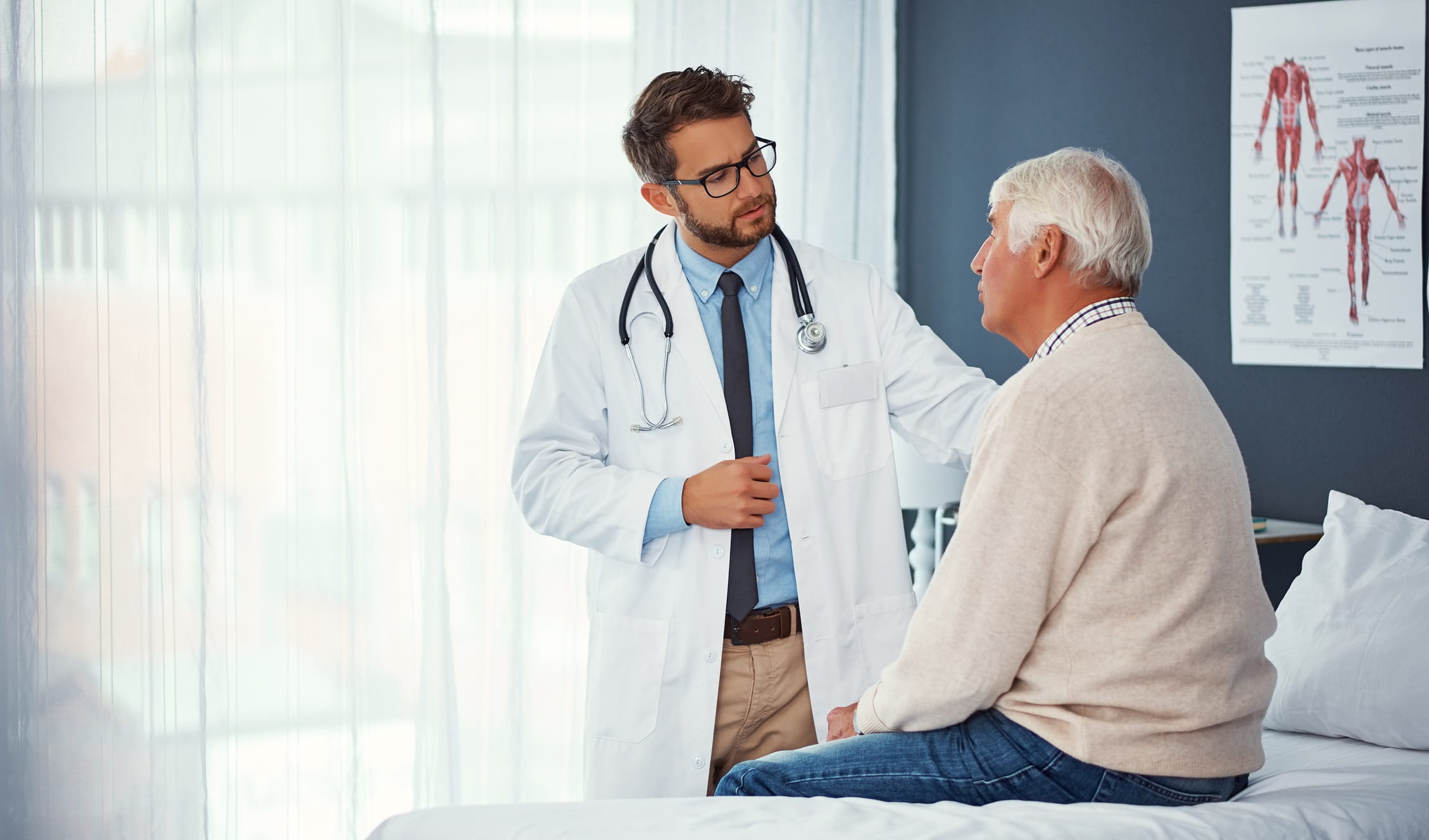Older man sitting in doctor's office with doctor standing nearby with hand on patient's shoulder Older man sitting in doctor's office with doctor standing nearby with hand on patient's shoulder