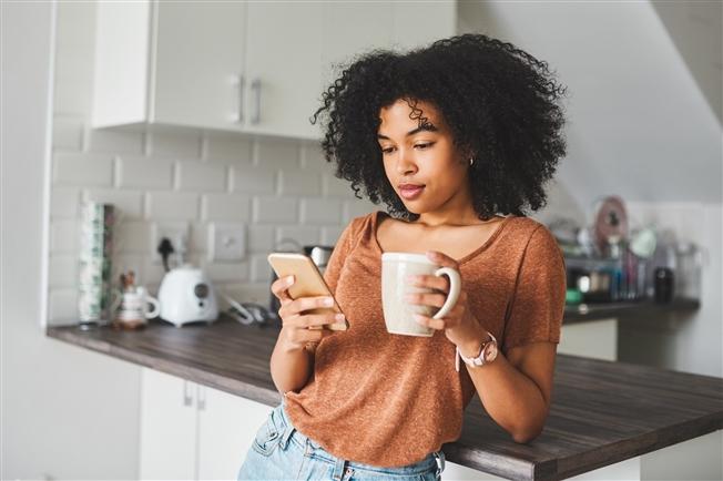 Woman leaning against kitchen counter, holding coffee cup and looking at smart phone Woman leaning against kitchen counter, holding coffee cup and looking at smart phone