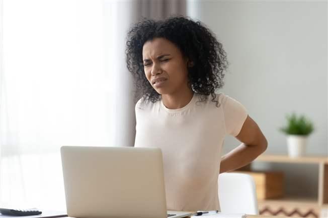 Young woman wincing at back pain from sitting at kitchen table and working Young woman wincing at back pain from sitting at kitchen table and working