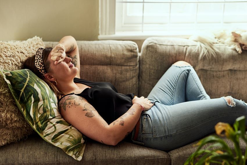 Young woman laying on couch grimacing in pain with hand to stomach