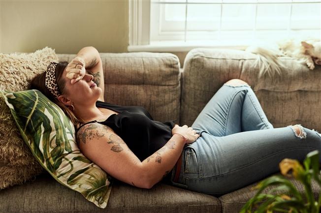 Young woman laying on couch grimacing in pain with hand to stomach Young woman laying on couch grimacing in pain with hand to stomach