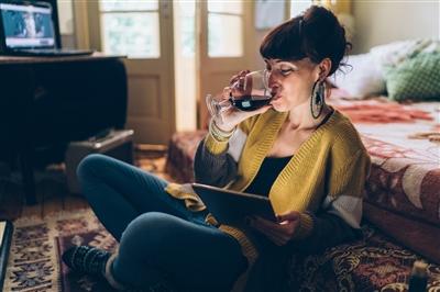 Young woman drinking glass of wine and watching TV show on tablet