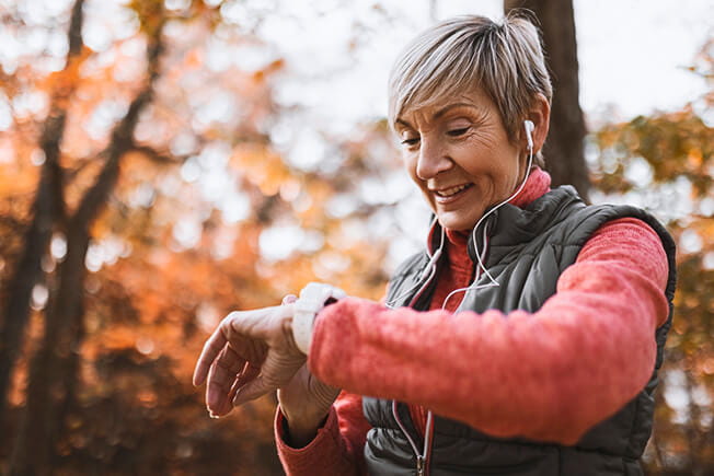 An older woman in the woods looking at her smart watch.