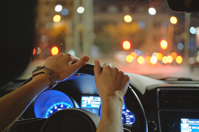 Close up of driver hands holding steering wheel driving car with blurred city street lights on background at night