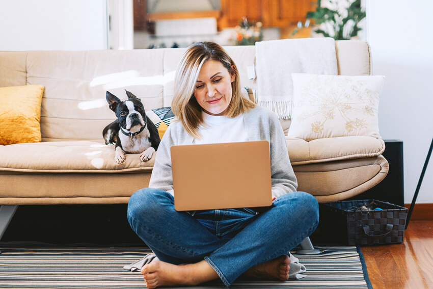 Woman sitting on the floor on her laptop with a dog on the couch behind her.