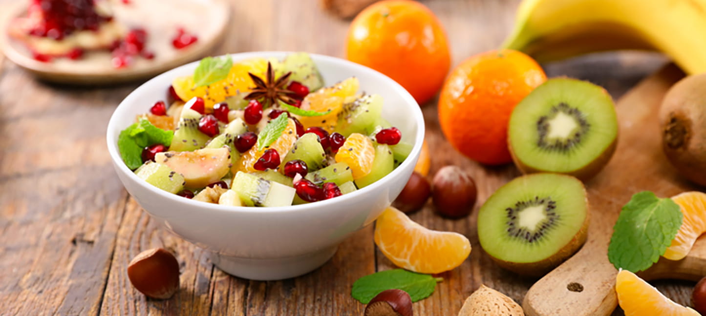Fruit salad in a bowl on a table with mixed fruit around the bowl.
