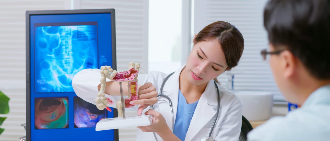 Young female Asian doctor explaining colon model to sick male patient at clinic with an x-ray film display on the computer behind her.