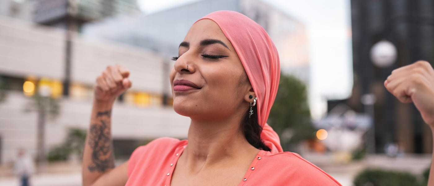 Young adult woman with pink cancer head scarf raising her arms flexing muscles.