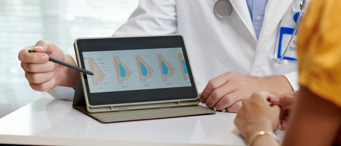 A doctor in a white coat shows a patient breast implant diagrams on a tablet.
