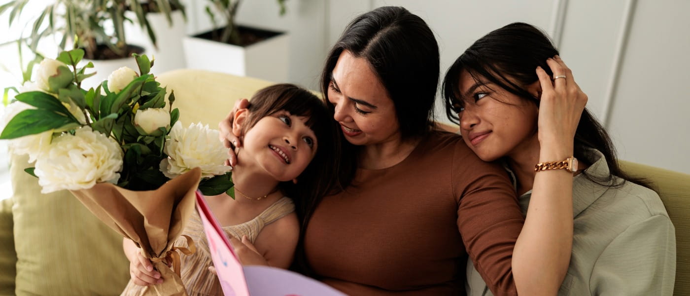 A mother sitting on a sofa with her two daughters, holding a bouquet of flowers and a handmade Mother’s Day card.