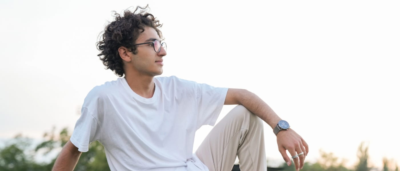 A young man with curly hair looking relaxed and thoughtful as he sits on the grass in an outdoor park.