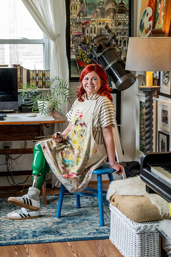 Kaitlin Gregory in an artist's smock, holding two paint brushes and seated on stool with her prosthetic legs crossed in her home.