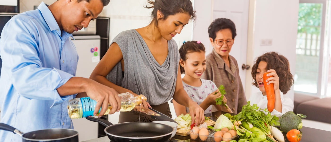 A dad pouring oil into a cooking pan, a mom chopping vegetables, a young girl and young boy examining the produce, and a grandmother observing from behind.