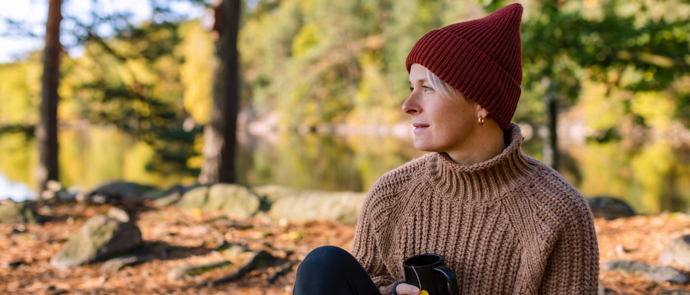 A woman in a sweater and knit cap sitting in front of a lake, under fir trees on a fall day while holding a mug of tea.