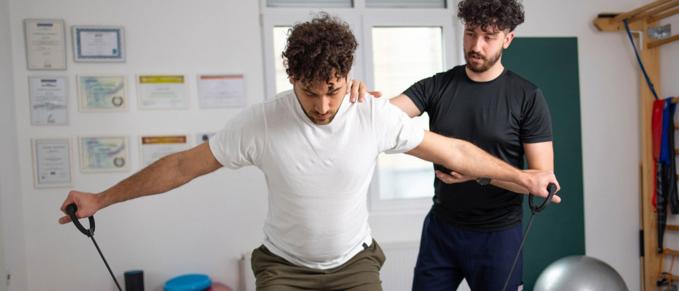 A man in a squat pulling at resistance bands in a workout room while a male physiotherapist stands behind, holding his shoulder to correct his balance.