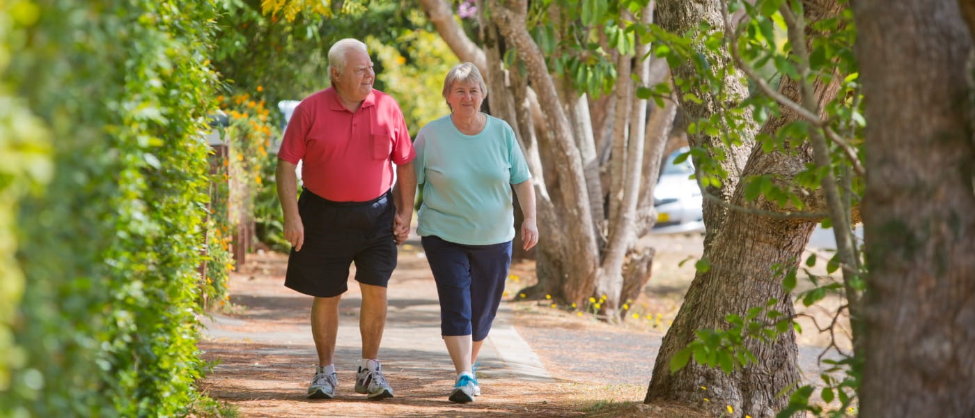 An older man holds the hand of an older woman as they walk down the sidewalk surrounded by greenery on a fence and leafy trees.