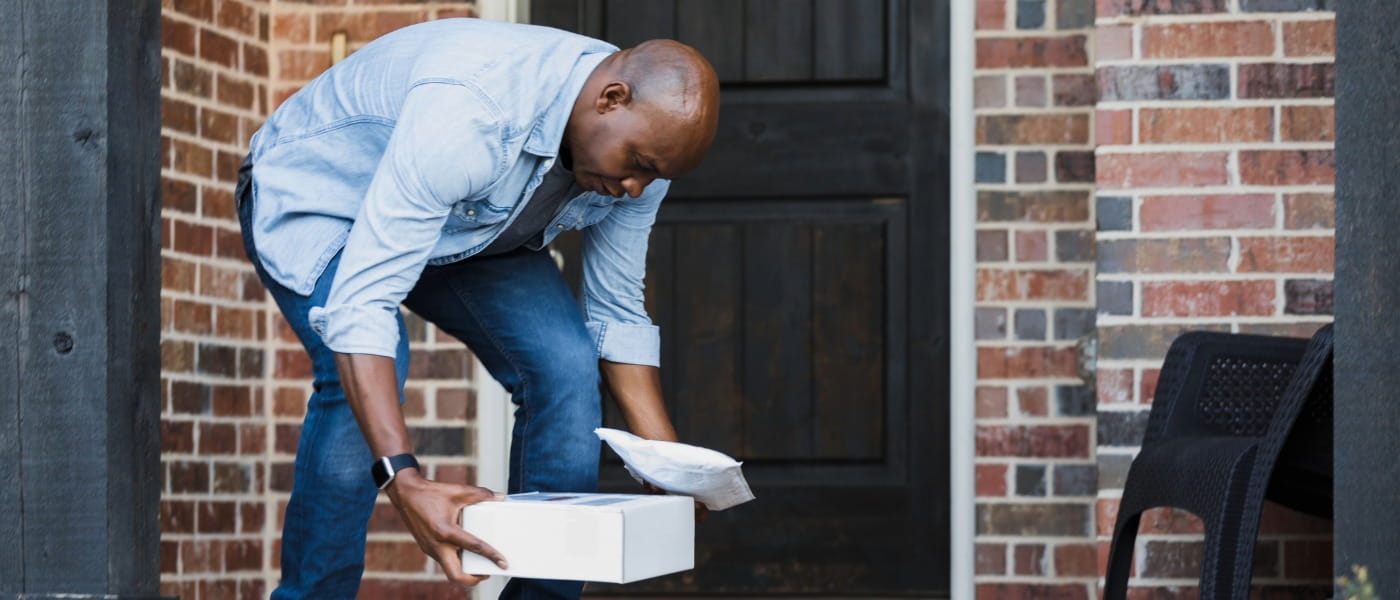 Mid-adult man bending down to pick up packages from the front porch of his home.