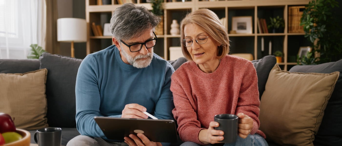 Senior man showing a senior woman results on a digital tablet as both have intrigued looks on their faces while they sit on their sofa with coffee mugs.