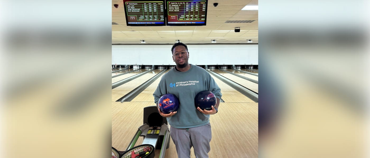 Bryce Ingram, MD holding two bowling balls in front of a lane at a bowling alley and wearing a sweatshirt that says Children's Hospital of Philadelphia.