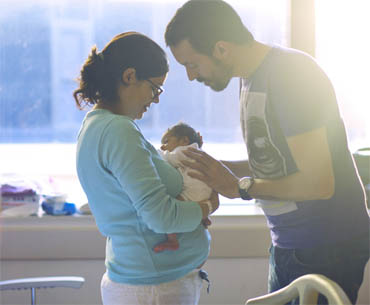 Mother holding newborn with father leans in over baby, all in hospital room