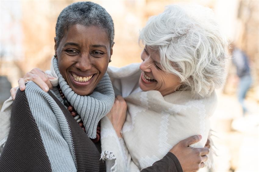 Two older women laughing and side hugging outside on a colder day