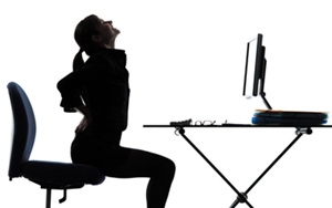 woman sitting on a chair in front of a computer desk cradling her pained lower back